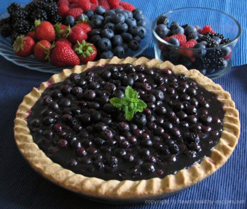 Whole blueberry pie topped with mint sprig and assortment of summer berries in the background.