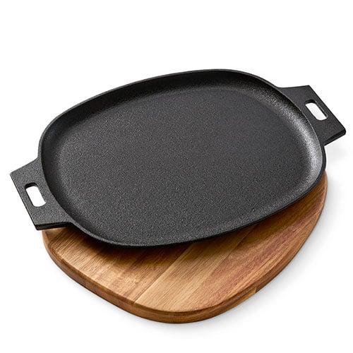 Black cast iron griddle with handles on a wooden trivet board against a white background.
