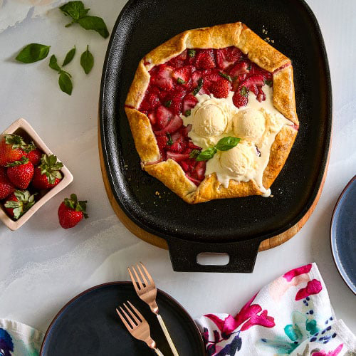 Strawberry galette with ice cream on a black cast iron skillet and wood trivet, surrounded by strawberries and a floral napkin.
