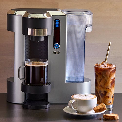 Deluxe Coffee Maker Machine with a cup of coffee, glass of iced coffee, and a plate with a cup of cappuccino on countertop surface.