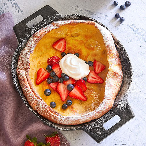 Dutch baby pancake with berries and whipped cream in medium-sized cast iron skillet on a textured surface.