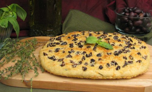 Whole, olive-basil focaccia bread garnished with basil, along with whole black olives and basil bunch in background.