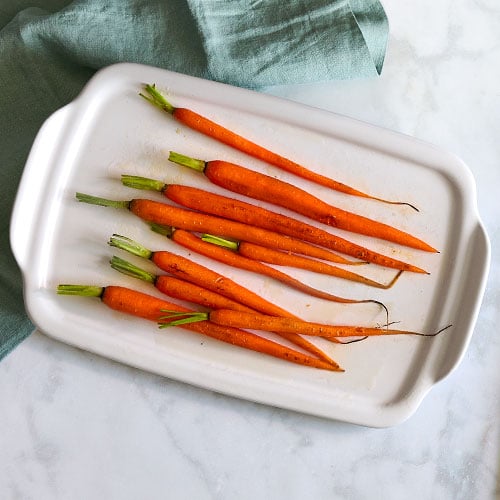 Cooked carrots on a stoneware serving and cooking tray placed on a marble countertop with greenish towel in background.