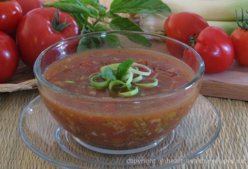 Tomato leek soup with garnish of chopped leek and basil sprig. Background of whole tomatoes, basil and leek stocks.