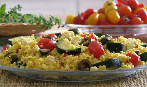 Greek-style couscous topped with tomatoes, zucchini and olives, along with herbs and tomatoes in background.