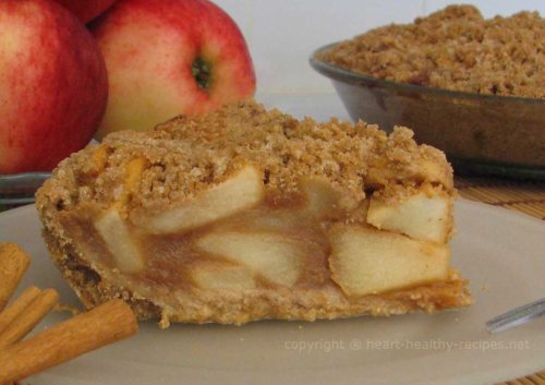 Close-up of apple pie with cinnamon sticks in foreground and bunch of apples and whole pie in background.