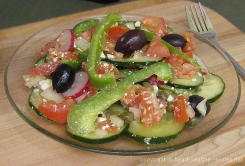 Greek salad on plate with black olives, green bell pepper slices, radish slices, tomatoes and cucumber slices.