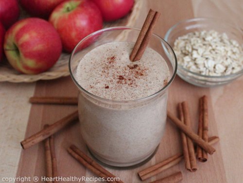 Apple cinnamon smoothie surrounded by cinnamon sticks along with whole apples and oats in the background.