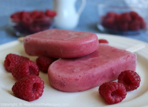 Two raspberry popsicles with several individual raspberries in foreground and more raspberries in background.
