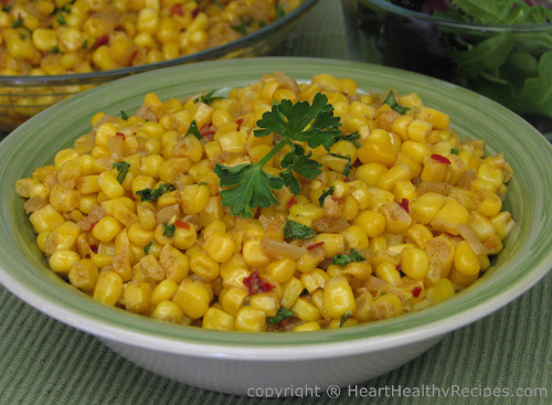 Corn sprinkled with green and red chilies topped with parsley along with more corn and a salad in background.