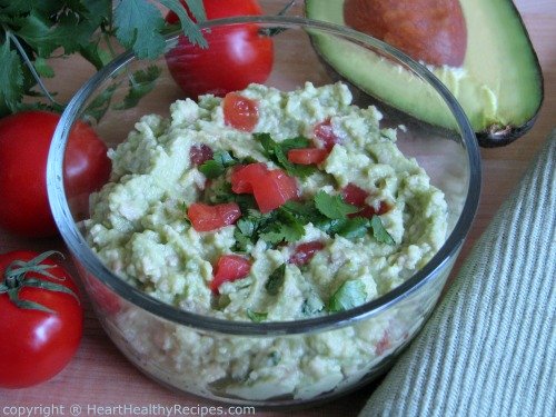 Fresh avocado guacamole with lime juice, onion, fresh cilantro, chopped tomatoes, garlic, surrounded by fresh tomatoes and halved avocado.