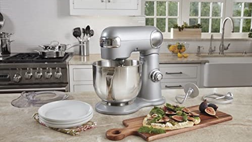 Silver colored, electric standing mixer with bowl displayed on kitchen countertop and surrounded by food.