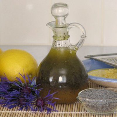 Honey Poppy Seed Dressing in glass container, along with whole lemons, poppy seeds, and purple flowers on kitchen countertop.