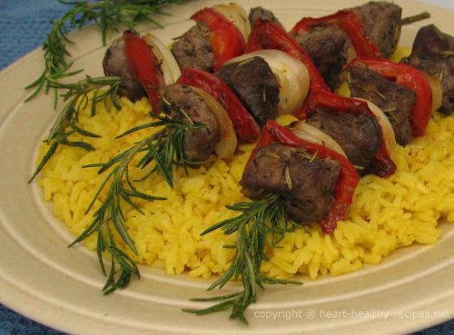 Rosemary lamb shish kabobs with red bell peppers and onion wedges on a rosemary sprig a top rice pilaf.