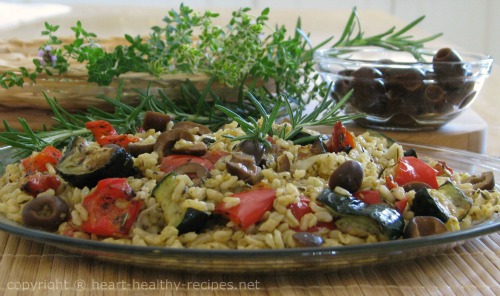 Roasted vegetable rice pilaf on serving plate with olives in bowl and herbs in background.